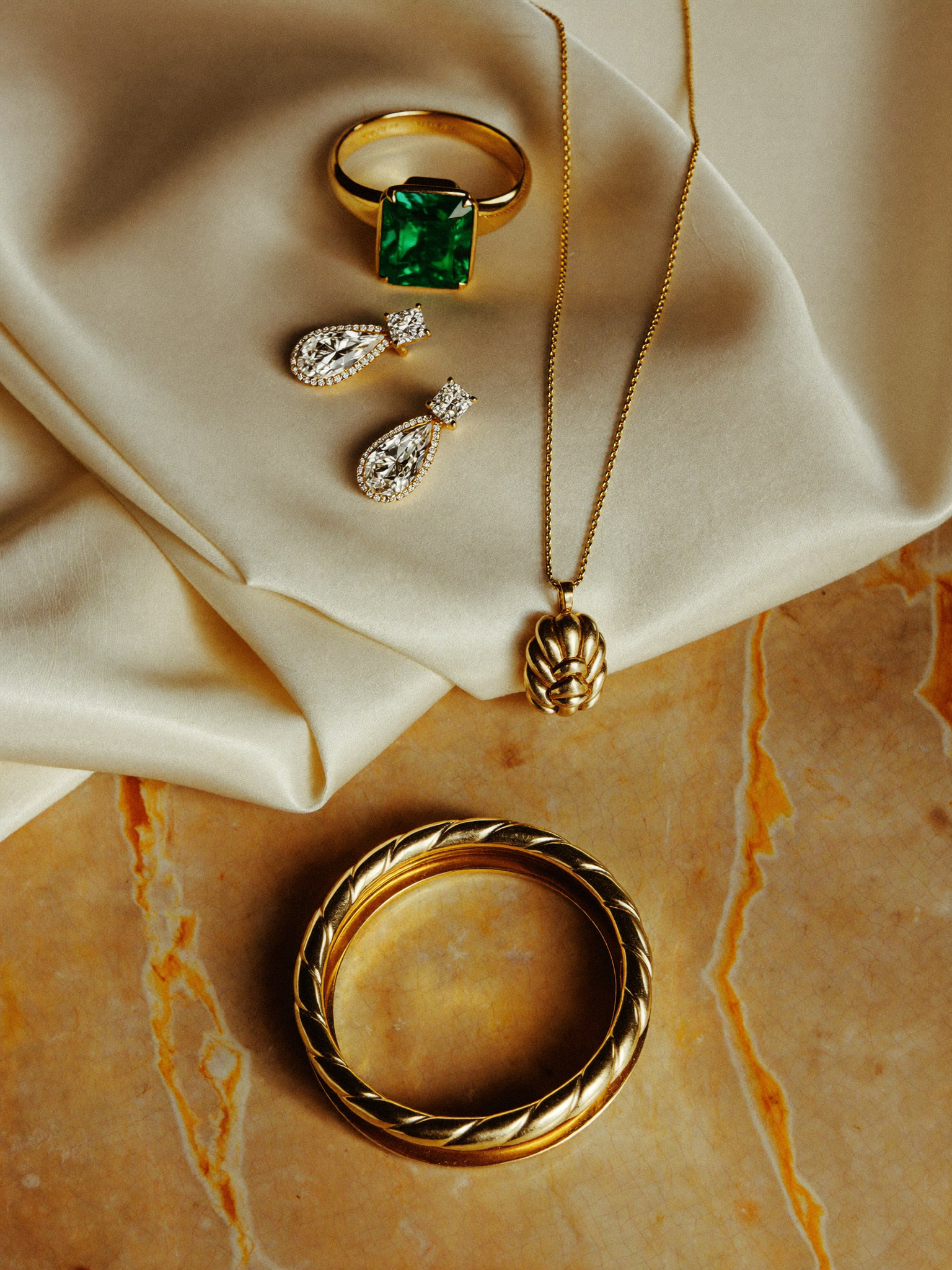 Flat-lay de joias JLópez — anel esmeralda, brincos, colar e pulseira sobre seda e mármore